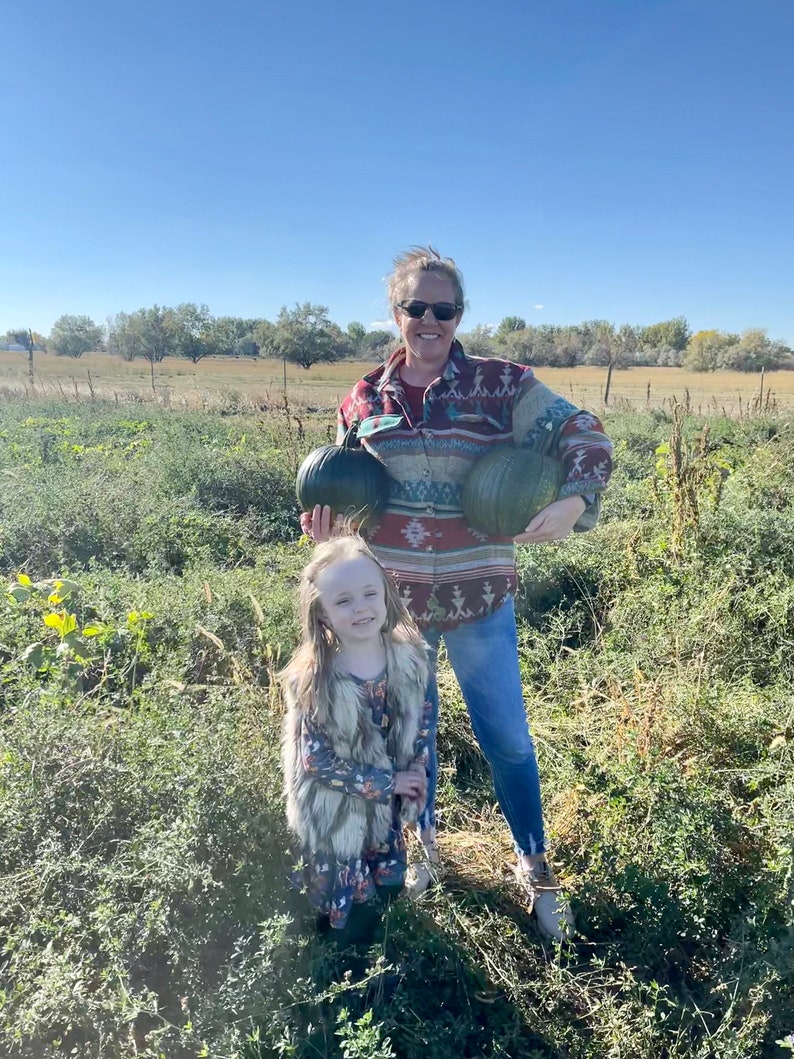 May include: A woman and a young girl are standing in a field, holding pumpkins. The woman is wearing a red and black patterned shirt, blue jeans, and sunglasses. The girl is wearing a brown faux fur vest over a floral dress.
