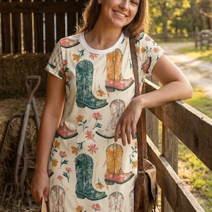 Può includere: Abito a maglietta color crema con un motivo ripetuto di stivali da cowboy in vari colori, tra cui verde, arancione e rosa, con accenti floreali. L'abito ha uno scollo bianco e maniche corte. Sono visibili anche un cappello di paglia e una borsa in pelle marrone.