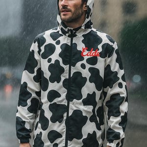 May include: A hooded jacket with a black and white cow print. The jacket has a zipper closure and a red "Edde" logo on the chest. The person is wearing the jacket in the rain.