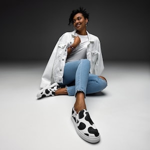 May include: Black and white cow print slip-on shoes with a white sole. The shoes are on a white surface. The person is wearing blue jeans and a white denim jacket.