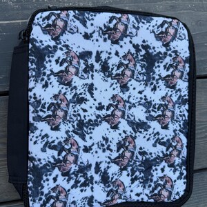 K&ouml;nnte beinhalten: Eine rechteckige Lunchtasche mit einem sich wiederholenden Muster aus braunen Pferden auf einem wei&szlig;-schwarzen Kuhfell-Hintergrund. Die Tasche verf&uuml;gt &uuml;ber einen schwarzen Rei&szlig;verschluss und eine schwarze Stoffeinfassung an den Kanten.