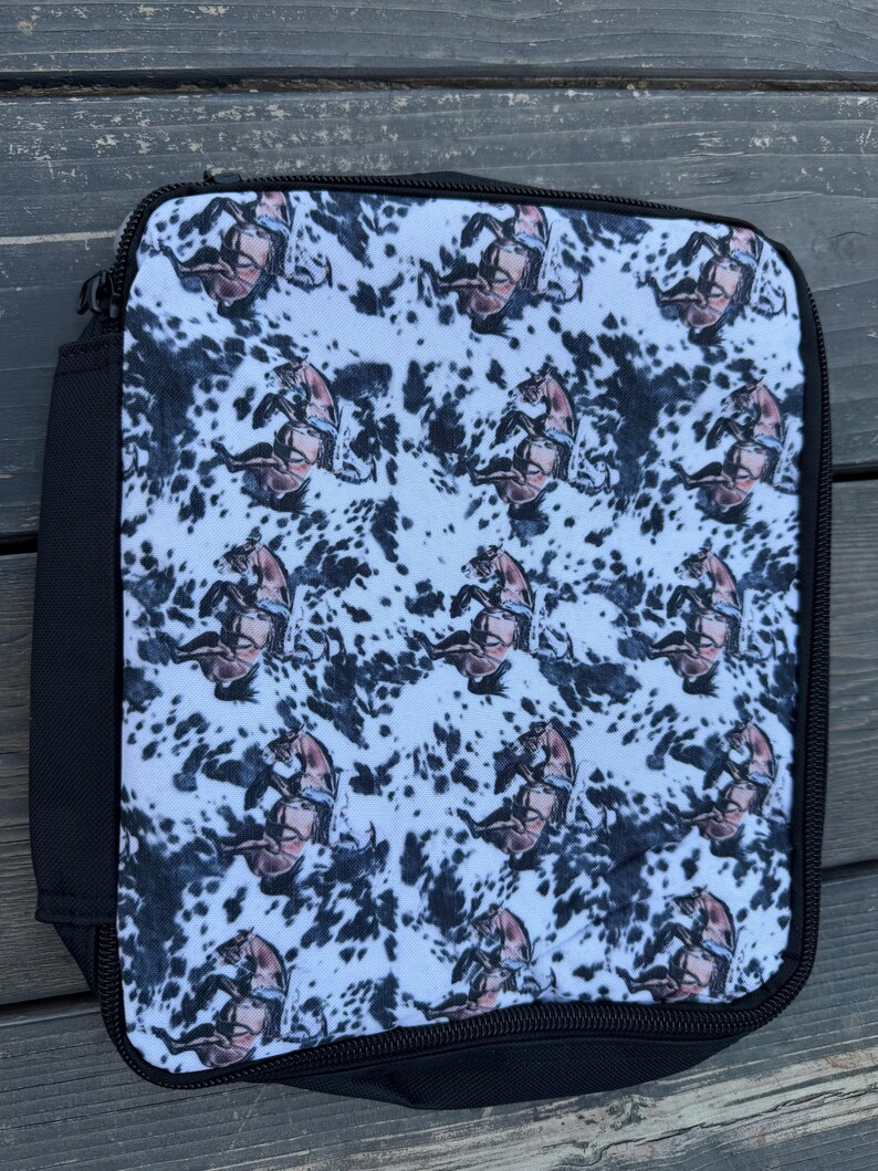 K&ouml;nnte beinhalten: Eine rechteckige Lunchtasche mit einem sich wiederholenden Muster aus braunen Pferden auf einem wei&szlig;-schwarzen Kuhfell-Hintergrund. Die Tasche verf&uuml;gt &uuml;ber einen schwarzen Rei&szlig;verschluss und eine schwarze Stoffeinfassung an den Kanten.