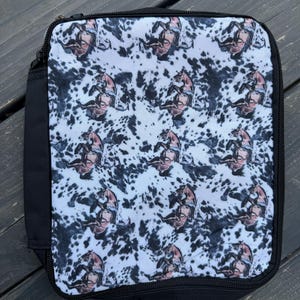 K&ouml;nnte beinhalten: Eine quadratische Lunchtasche mit schwarzem Rei&szlig;verschluss und schwarzen Seitenteilen. Die Vorderseite zeigt ein sich wiederholendes Muster aus braunen Pferden auf wei&szlig;em Grund mit schwarzen Flecken im Kuhfell-Stil.