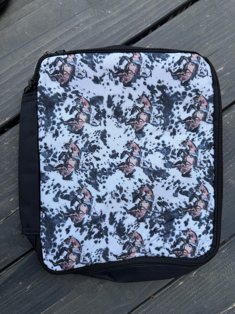 K&ouml;nnte beinhalten: Eine quadratische Lunchtasche mit schwarzem Rei&szlig;verschluss und schwarzen Seitenteilen. Die Vorderseite zeigt ein sich wiederholendes Muster aus braunen Pferden auf wei&szlig;em Grund mit schwarzen Flecken im Kuhfell-Stil.