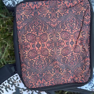 K&ouml;nnte beinhalten: Ein quadratisches Aufbewahrungsetui mit Rei&szlig;verschluss und einem sich wiederholenden Blumenmuster in gebranntem Orange und Schwarz. Das Etui hat schwarze Stoffr&auml;nder und einen schwarzen Rei&szlig;verschluss. Die Oberfl&auml;che wirkt weich und flexibel.