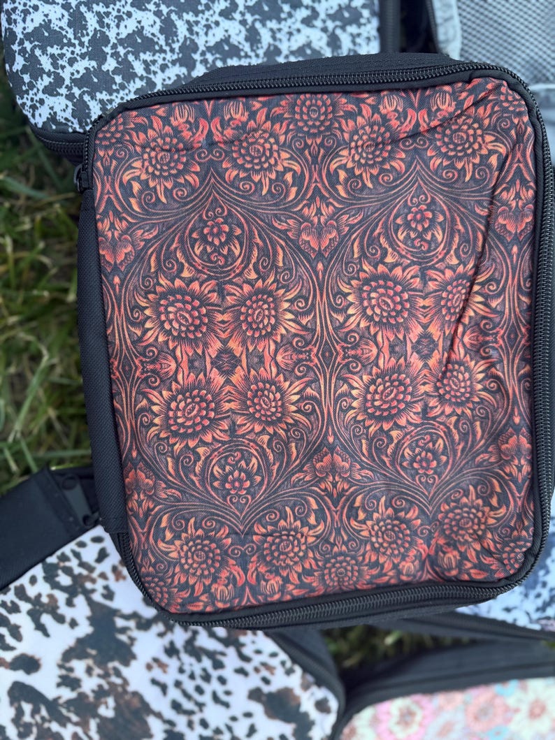 K&ouml;nnte beinhalten: Ein quadratisches Aufbewahrungsetui mit Rei&szlig;verschluss und einem sich wiederholenden Blumenmuster in gebranntem Orange und Schwarz. Das Etui hat schwarze Stoffr&auml;nder und einen schwarzen Rei&szlig;verschluss. Die Oberfl&auml;che wirkt weich und flexibel.