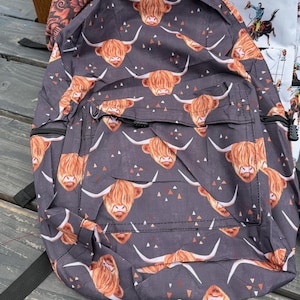 K&ouml;nnte beinhalten: Ein dunkelgrauer Rucksack mit einem Allover-Print aus braunen Hochlandrind-K&ouml;pfen mit langen H&ouml;rnern und kleinen verstreuten orangefarbenen und wei&szlig;en Dreiecken. Die Tasche verf&uuml;gt &uuml;ber eine Fronttasche mit schwarzen Rei&szlig;verschl&uuml;ssen und Riemen.