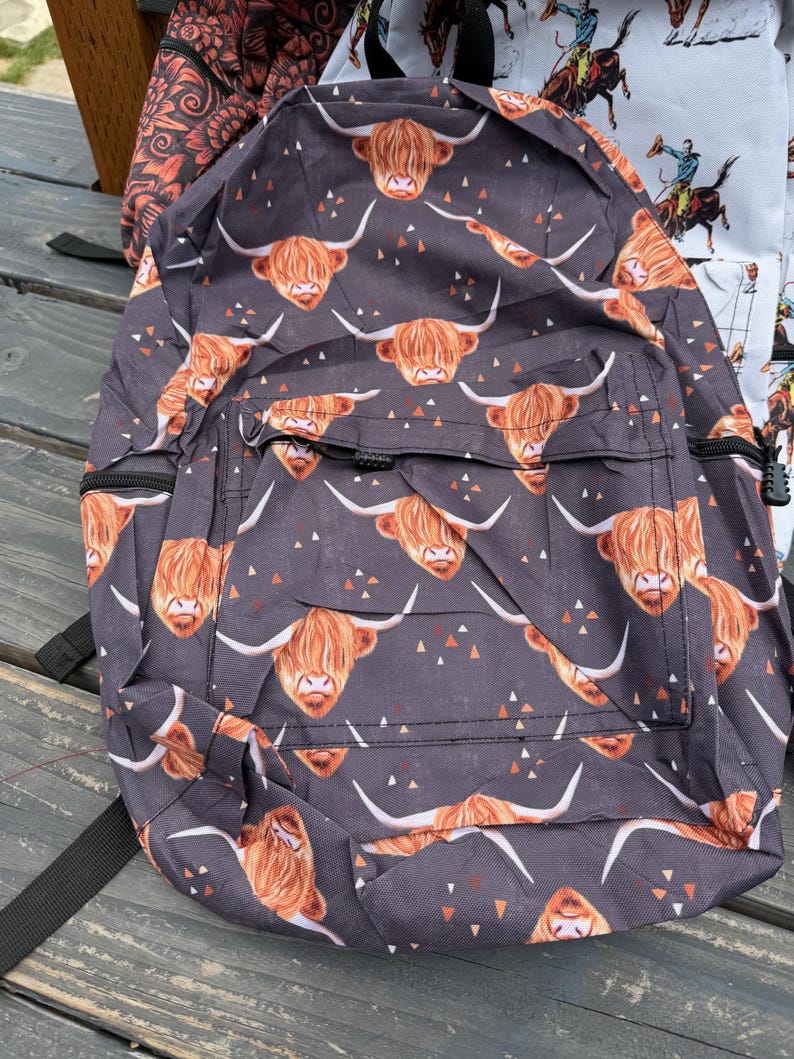 K&ouml;nnte beinhalten: Ein dunkelgrauer Rucksack mit einem Allover-Print aus braunen Hochlandrind-K&ouml;pfen mit langen H&ouml;rnern und kleinen verstreuten orangefarbenen und wei&szlig;en Dreiecken. Die Tasche verf&uuml;gt &uuml;ber eine Fronttasche mit schwarzen Rei&szlig;verschl&uuml;ssen und Riemen.