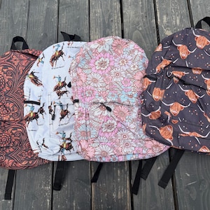 K&ouml;nnte beinhalten: Vier Rucks&auml;cke mit verschiedenen Mustern: einer mit dunkelorangem Blumenmuster, einer mit wei&szlig;em Cowboy- und Pferdemotiv, einer mit rosa-wei&szlig;em Blumendesign und einer mit schwarzem Hintergrund und braunen Highland-Rind-Illustrationen.