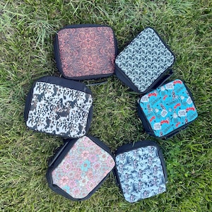 K&ouml;nnte beinhalten: Sechs quadratische Lunchtaschen mit schwarzem Rand und Rei&szlig;verschl&uuml;ssen, jede mit einem eigenen Muster: ein brauner Blumendruck, ein schwarz-wei&szlig;es Kuhfellmuster, ein rosa-blaues Blumendesign, ein schwarz-wei&szlig; gesprenkeltes Muster, ein blau-wei&szlig;er Batik-Effekt und ein blauer Aufdruck mit Cowboy-Motiven.