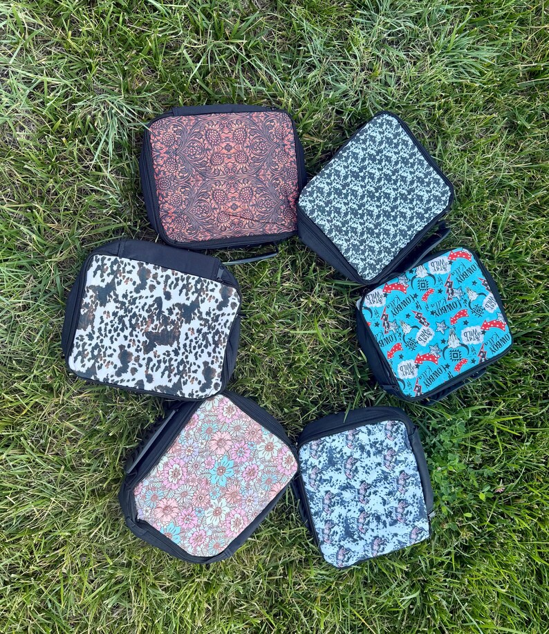 K&ouml;nnte beinhalten: Sechs quadratische Lunchtaschen mit schwarzem Rand und Rei&szlig;verschl&uuml;ssen, jede mit einem eigenen Muster: ein brauner Blumendruck, ein schwarz-wei&szlig;es Kuhfellmuster, ein rosa-blaues Blumendesign, ein schwarz-wei&szlig; gesprenkeltes Muster, ein blau-wei&szlig;er Batik-Effekt und ein blauer Aufdruck mit Cowboy-Motiven.