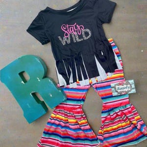 Stay Wild Girls Bell Bottom Set. Features a black Fringed Shirt with a bold "Stay Wild" design in pink and cheetah print, perfectly paired with vibrant serape-striped bell bottoms. Ships FREE with $35+ purchase.