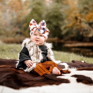 May include: A baby wearing a brown and white faux fur vest, a black long-sleeved shirt, and brown and white striped pants. The baby is wearing a colorful bow headband and is sitting on a brown and white cowhide rug.