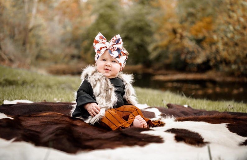May include: A baby wearing a brown and white faux fur vest, a black long-sleeved shirt, and brown and white striped pants. The baby is wearing a colorful bow headband and is sitting on a brown and white cowhide rug.