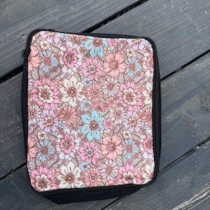 K&ouml;nnte beinhalten: Eine rechteckige Rei&szlig;verschlusstasche mit einem dichten Blumenmuster aus rosa, hellblauen und wei&szlig;en Bl&uuml;ten auf cremefarbenem Grund. Die Tasche hat einen einfarbigen schwarzen Stoffrand und einen schwarzen Rei&szlig;verschluss an der Ober- und Seite.