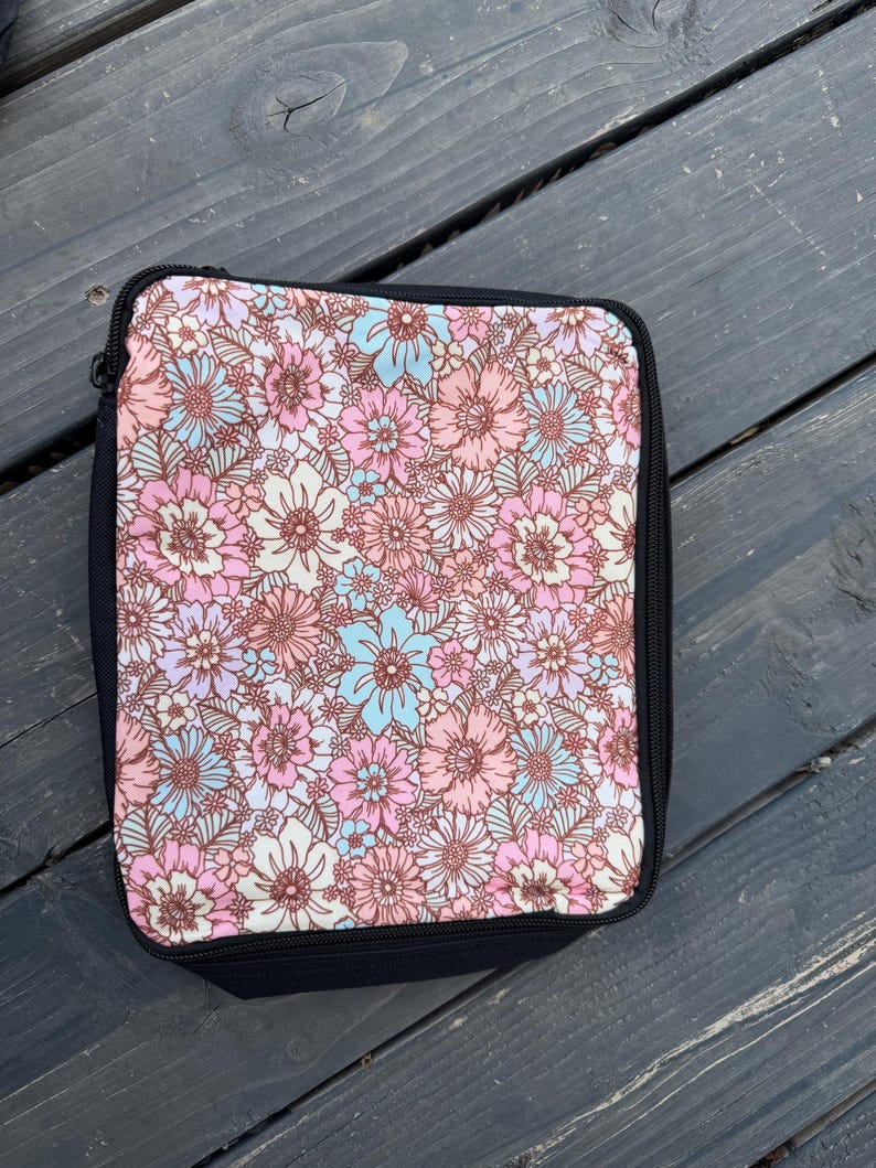 K&ouml;nnte beinhalten: Eine rechteckige Rei&szlig;verschlusstasche mit einem dichten Blumenmuster aus rosa, hellblauen und wei&szlig;en Bl&uuml;ten auf cremefarbenem Grund. Die Tasche hat einen einfarbigen schwarzen Stoffrand und einen schwarzen Rei&szlig;verschluss an der Ober- und Seite.