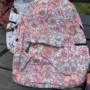K&ouml;nnte beinhalten: Ein Rucksack mit einem sich wiederholenden Blumenmuster in Rosa-, Hellblau- und Wei&szlig;t&ouml;nen. Die Tasche verf&uuml;gt &uuml;ber eine vordere Rei&szlig;verschlusstasche mit schwarzem Zipper und ein Hauptfach, das eine dichte Anordnung verschiedener Blumen zeigt.