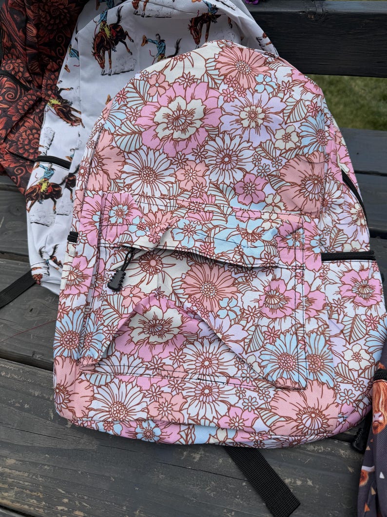 K&ouml;nnte beinhalten: Ein Rucksack mit einem sich wiederholenden Blumenmuster in Rosa-, Hellblau- und Wei&szlig;t&ouml;nen. Die Tasche verf&uuml;gt &uuml;ber eine vordere Rei&szlig;verschlusstasche mit schwarzem Zipper und ein Hauptfach, das eine dichte Anordnung verschiedener Blumen zeigt.