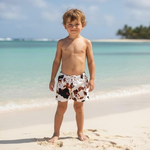 Peut inclure: Un jeune enfant se tient sur une plage de sable, portant un short de bain blanc avec un motif de peau de vache marron et noir. Le short a une fermeture à cordon. L'arrière-plan présente un océan turquoise et un ciel bleu.