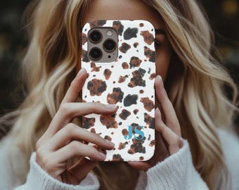 May include: A white phone case with a brown and black cow print pattern. The case has a blue monogram of the letters "JS" at the bottom. The phone case is held in front of a person.