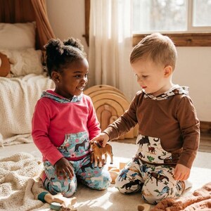 Puede incluir: Dos ni&ntilde;os con conjuntos a juego juegan con juguetes de madera. Los conjuntos incluyen una sudadera con capucha rosa y marr&oacute;n con un bolsillo con estampado de caballos y pantalones a juego. Los ni&ntilde;os est&aacute;n sentados en una alfombra color crema.