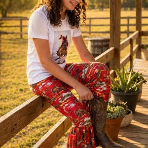 Può includere: T-shirt bianco con grafica di cowboy e pantaloni rossi con stampa vintage di cowboy. I pantaloni hanno un fondo svasato. L'immagine mostra anche stivali da cowboy marroni. Il testo "VINTAGE COWBOY PRINT" è in alto.