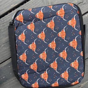 K&ouml;nnte beinhalten: Eine rechteckige Lunchtasche mit dunkelblauem Hintergrund und einem wiederkehrenden Muster aus orangefarbenen und braunen Hochlandrind-K&ouml;pfen mit langen wei&szlig;en H&ouml;rnern und kleinen orangefarbenen Dreiecken, versehen mit einem schwarzen Rei&szlig;verschluss.