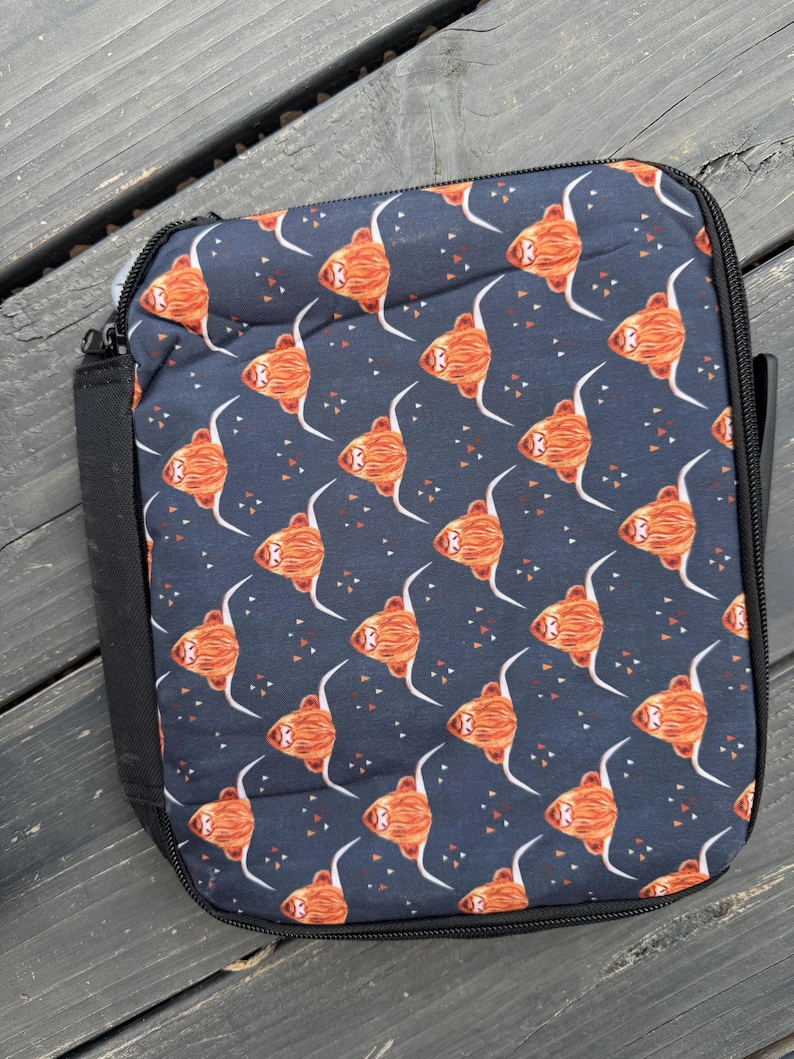 K&ouml;nnte beinhalten: Eine rechteckige Lunchtasche mit dunkelblauem Hintergrund und einem wiederkehrenden Muster aus orangefarbenen und braunen Hochlandrind-K&ouml;pfen mit langen wei&szlig;en H&ouml;rnern und kleinen orangefarbenen Dreiecken, versehen mit einem schwarzen Rei&szlig;verschluss.