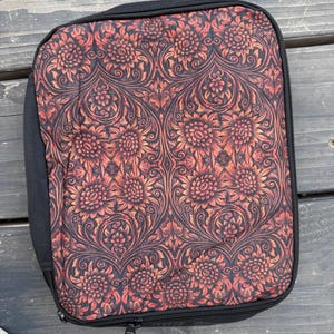 K&ouml;nnte beinhalten: Ein rechteckiges Etui mit Rei&szlig;verschluss und einem floralen Damastmuster in Schwarz und gebranntem Orange. Das Etui hat einen schwarzen Stoffrand und einen schwarzen Rei&szlig;verschluss, der entlang des Umfangs verl&auml;uft und den Inhalt sicher umschlie&szlig;t.