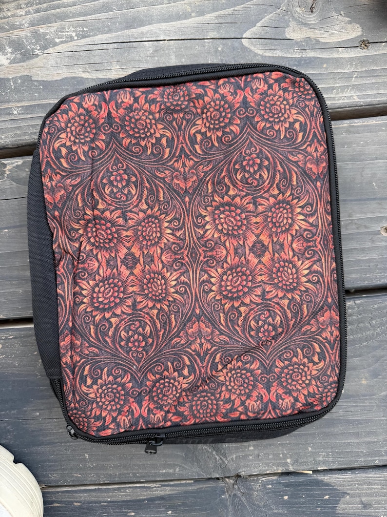 K&ouml;nnte beinhalten: Ein rechteckiges Etui mit Rei&szlig;verschluss und einem floralen Damastmuster in Schwarz und gebranntem Orange. Das Etui hat einen schwarzen Stoffrand und einen schwarzen Rei&szlig;verschluss, der entlang des Umfangs verl&auml;uft und den Inhalt sicher umschlie&szlig;t.