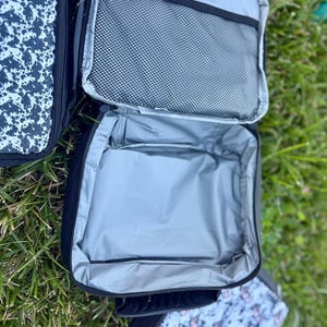 K&ouml;nnte beinhalten: Eine rechteckige Lunchtasche mit schwarzer Au&szlig;enseite und silberner, isolierter Innenauskleidung. Die Tasche verf&uuml;gt &uuml;ber ein Hauptfach mit Rei&szlig;verschluss und eine schwarze Netztasche an der Innenseite des Deckels. Zwei weitere Taschen mit schwarz-wei&szlig;em Muster sind zu sehen.
