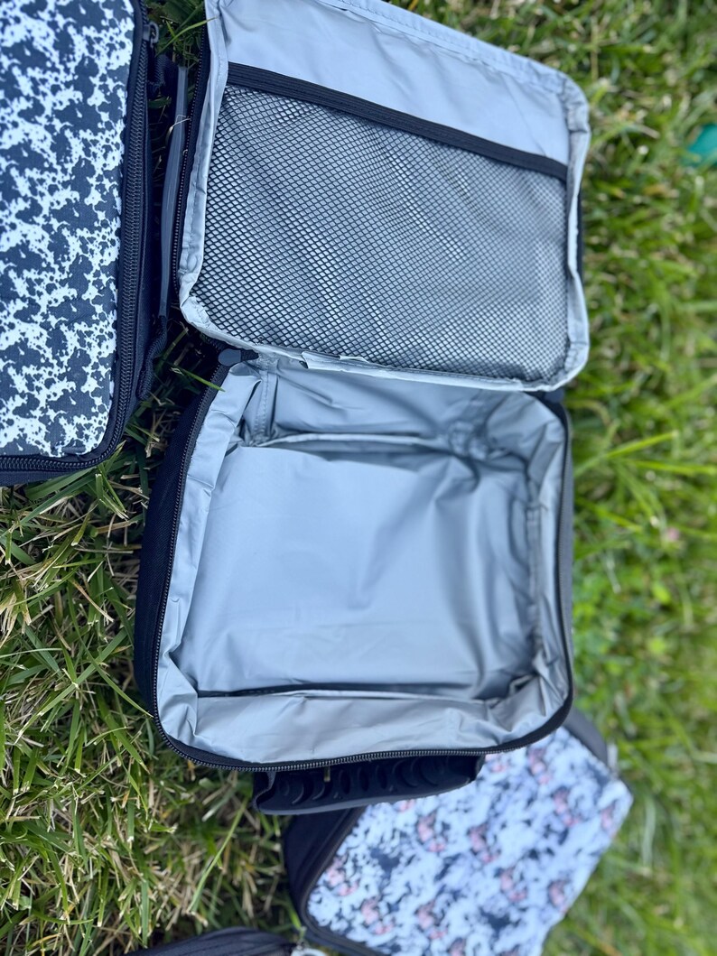 K&ouml;nnte beinhalten: Eine rechteckige Lunchtasche mit schwarzer Au&szlig;enseite und silberner, isolierter Innenauskleidung. Die Tasche verf&uuml;gt &uuml;ber ein Hauptfach mit Rei&szlig;verschluss und eine schwarze Netztasche an der Innenseite des Deckels. Zwei weitere Taschen mit schwarz-wei&szlig;em Muster sind zu sehen.