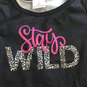 Stay Wild Girls Bell Bottom Set. Features a black Fringed Shirt with a bold "Stay Wild" design in pink and cheetah print, perfectly paired with vibrant serape-striped bell bottoms. Ships FREE with $35+ purchase.