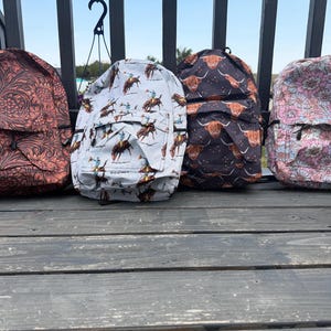 K&ouml;nnte beinhalten: Vier Rucks&auml;cke in einer Reihe. Der erste hat ein dunkelbraunes Blumenmuster, der zweite einen hellblauen Cowboy-Druck, der dritte ein schwarzes Hochlandrind-Muster und der vierte einen hellrosa-wei&szlig;en Blumendruck. Jeder hat eine Vordertasche.