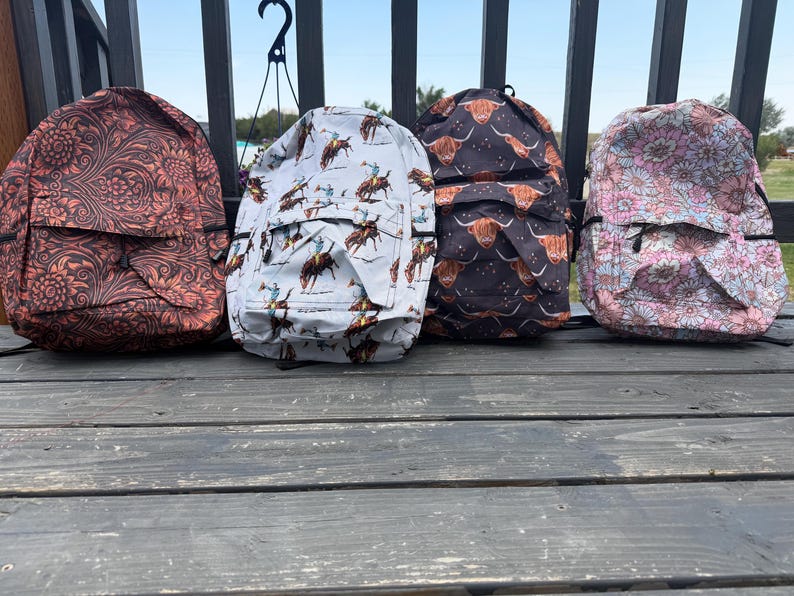 K&ouml;nnte beinhalten: Vier Rucks&auml;cke in einer Reihe. Der erste hat ein dunkelbraunes Blumenmuster, der zweite einen hellblauen Cowboy-Druck, der dritte ein schwarzes Hochlandrind-Muster und der vierte einen hellrosa-wei&szlig;en Blumendruck. Jeder hat eine Vordertasche.