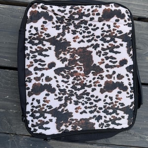 K&ouml;nnte beinhalten: Eine rechteckige Tasche mit Rei&szlig;verschluss, die auf der Vorderseite ein wei&szlig;es, schwarzes und braunes Kuhfellmuster aufweist. Die Tasche ist von einem schwarzen Stoffrand umgeben und hat einen schwarzen Rei&szlig;verschluss entlang des Randes.