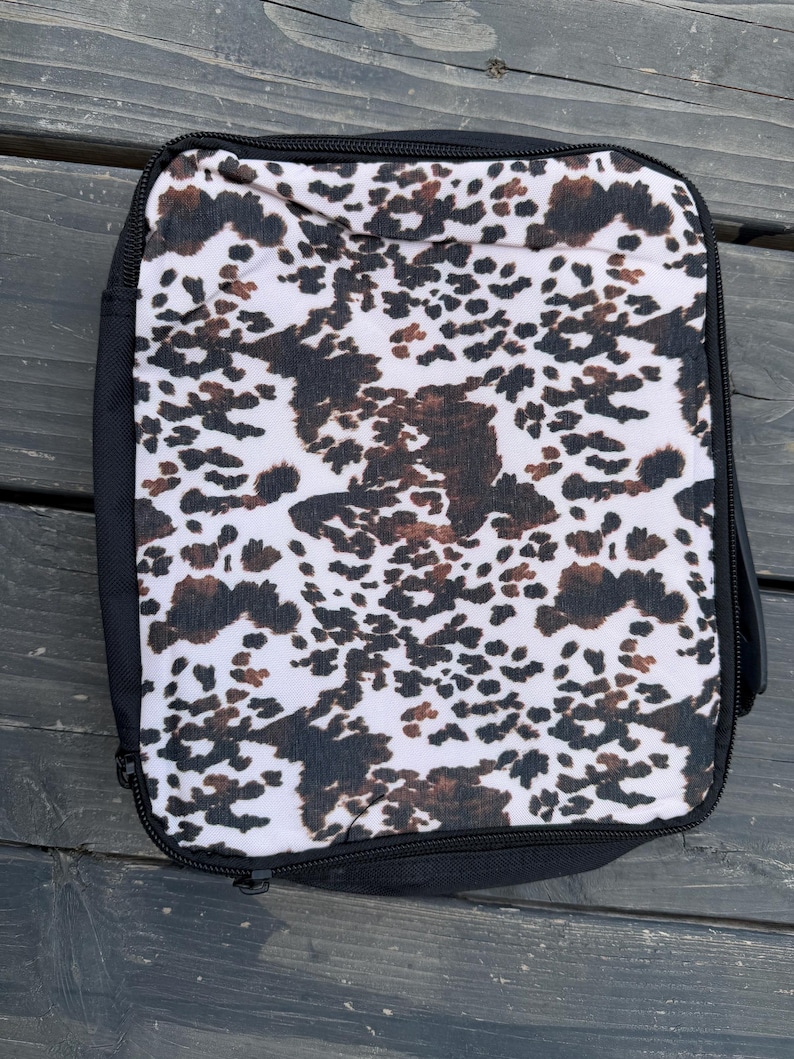 K&ouml;nnte beinhalten: Eine rechteckige Tasche mit Rei&szlig;verschluss, die auf der Vorderseite ein wei&szlig;es, schwarzes und braunes Kuhfellmuster aufweist. Die Tasche ist von einem schwarzen Stoffrand umgeben und hat einen schwarzen Rei&szlig;verschluss entlang des Randes.
