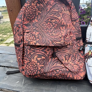 K&ouml;nnte beinhalten: Ein Rucksack mit einem dunkelbraunen und orangefarbenen Blumenmuster, ausgestattet mit einem gro&szlig;en Hauptfach und einer kleineren Vordertasche. Das Design besteht aus komplizierten, sich wiederholenden Blumen- und Rankenmotiven.