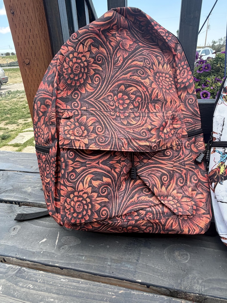 K&ouml;nnte beinhalten: Ein Rucksack mit einem dunkelbraunen und orangefarbenen Blumenmuster, ausgestattet mit einem gro&szlig;en Hauptfach und einer kleineren Vordertasche. Das Design besteht aus komplizierten, sich wiederholenden Blumen- und Rankenmotiven.