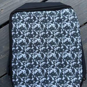 K&ouml;nnte beinhalten: Ein rechteckiges Etui mit Rei&szlig;verschluss, das auf der Vorderseite ein schwarz-wei&szlig;es Sprenkelmuster aufweist. Das Etui ist mit einfarbigen schwarzen Stoffr&auml;ndern und einem schwarzen Rei&szlig;verschluss versehen, der entlang des rechteckigen Umrisses verl&auml;uft.