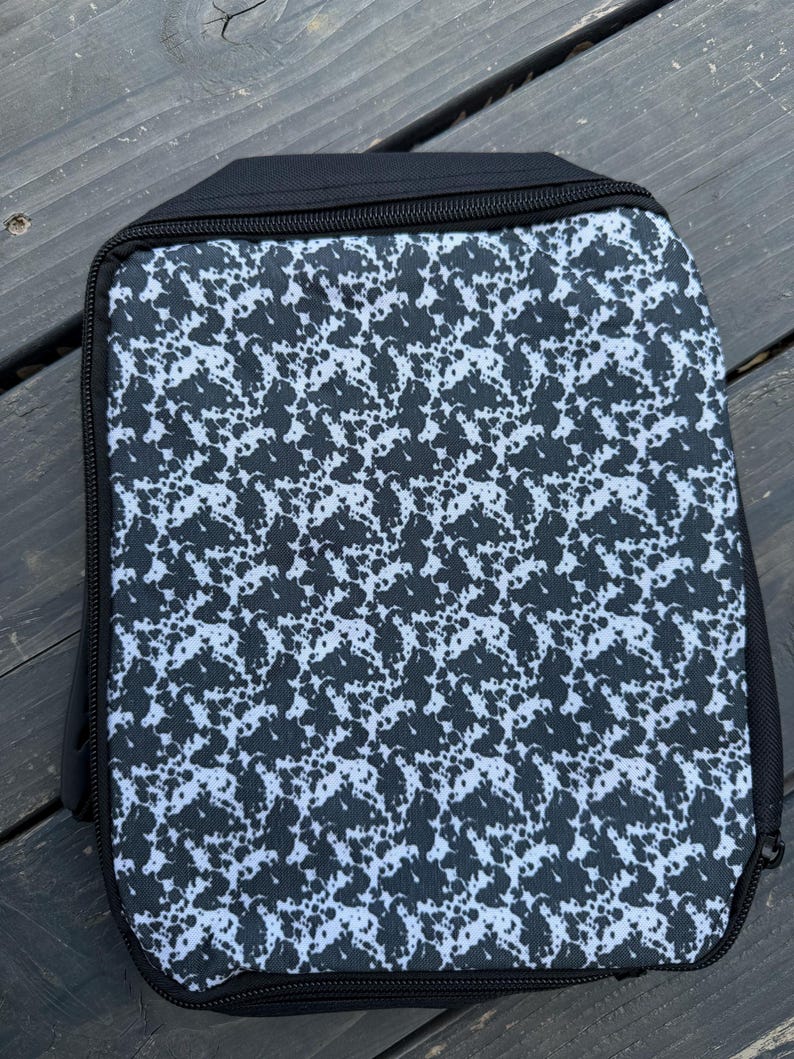 K&ouml;nnte beinhalten: Ein rechteckiges Etui mit Rei&szlig;verschluss, das auf der Vorderseite ein schwarz-wei&szlig;es Sprenkelmuster aufweist. Das Etui ist mit einfarbigen schwarzen Stoffr&auml;ndern und einem schwarzen Rei&szlig;verschluss versehen, der entlang des rechteckigen Umrisses verl&auml;uft.