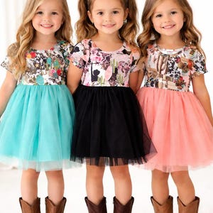 May include: Three young girls wearing matching dresses with floral tops and tulle skirts in turquoise, black, and coral. They are wearing brown cowboy boots. The dresses have a Western theme.
