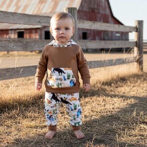 Puede incluir: Un ni&ntilde;o peque&ntilde;o con una sudadera con capucha marr&oacute;n y pantalones a juego con un estampado de tem&aacute;tica occidental. La sudadera tiene un bolsillo estampado y forro de capucha. Los pantalones presentan un patr&oacute;n repetido de vaqueros, caballos y cactus.