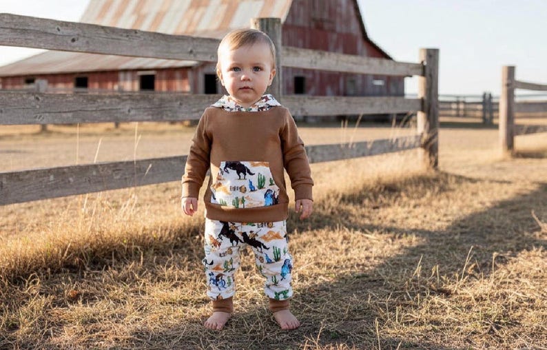 Puede incluir: Un ni&ntilde;o peque&ntilde;o con una sudadera con capucha marr&oacute;n y pantalones a juego con un estampado de tem&aacute;tica occidental. La sudadera tiene un bolsillo estampado y forro de capucha. Los pantalones presentan un patr&oacute;n repetido de vaqueros, caballos y cactus.