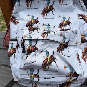 K&ouml;nnte beinhalten: Ein wei&szlig;er Rucksack mit einem sich wiederholenden Muster aus Cowboys auf bockenden Pferden. Der Druck zeigt Figuren in blauen Hemden und gelben Hosen auf braunen Pferden, wobei jeder Reiter einen Hut in die Luft h&auml;lt. Mit einer Fronttasche mit Rei&szlig;verschluss.