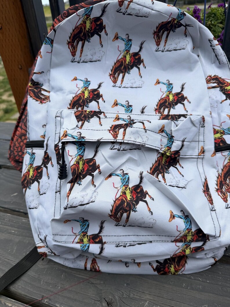 K&ouml;nnte beinhalten: Ein wei&szlig;er Rucksack mit einem sich wiederholenden Muster aus Cowboys auf bockenden Pferden. Der Druck zeigt Figuren in blauen Hemden und gelben Hosen auf braunen Pferden, wobei jeder Reiter einen Hut in die Luft h&auml;lt. Mit einer Fronttasche mit Rei&szlig;verschluss.