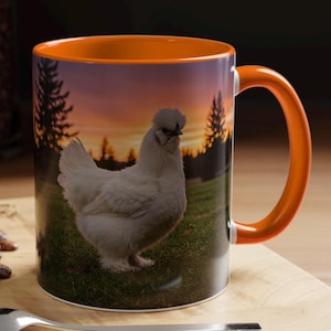 May include: A white Silkie chicken with a fluffy crest stands in a grassy field at sunset. The chicken is facing the right side of the image. The background is a blurry sunset with trees in the distance. The image is printed on a white ceramic mug with an orange rim and handle.