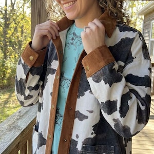 May include: A button-down jacket featuring a black and white cow print pattern, accented with a brown corduroy collar and sleeve cuffs. The jacket includes gold-toned metal buttons and two front patch pockets, worn over a light blue t-shirt.