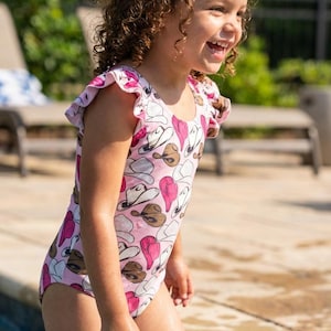 K&ouml;nnte beinhalten: Ein rosa Badeanzug mit Cowboyhut-Muster. Der Badeanzug hat R&uuml;schen&auml;rmel und wird von einer jungen Person in einem Pool getragen. Der Hintergrund umfasst einen Pool, eine Terrasse und Gr&uuml;n.