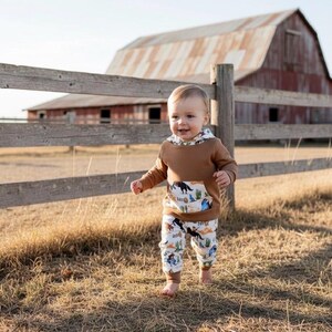 Puede incluir: Un ni&ntilde;o peque&ntilde;o con una sudadera con capucha marr&oacute;n con un bolsillo con una escena del desierto y pantalones a juego. El conjunto se encuentra frente a una valla de madera r&uacute;stica y un granero.